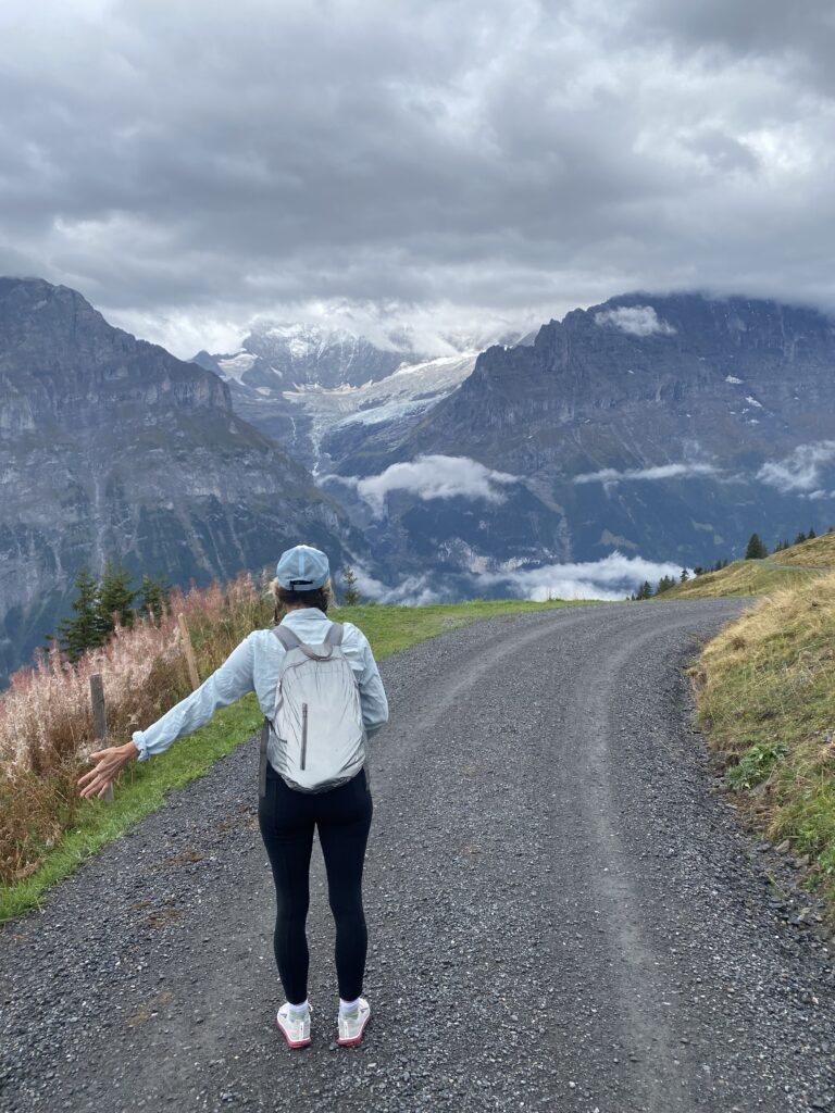 Woman walking on a mountain path in the Swiss Alps surrounded by clouds and dramatic peaks.