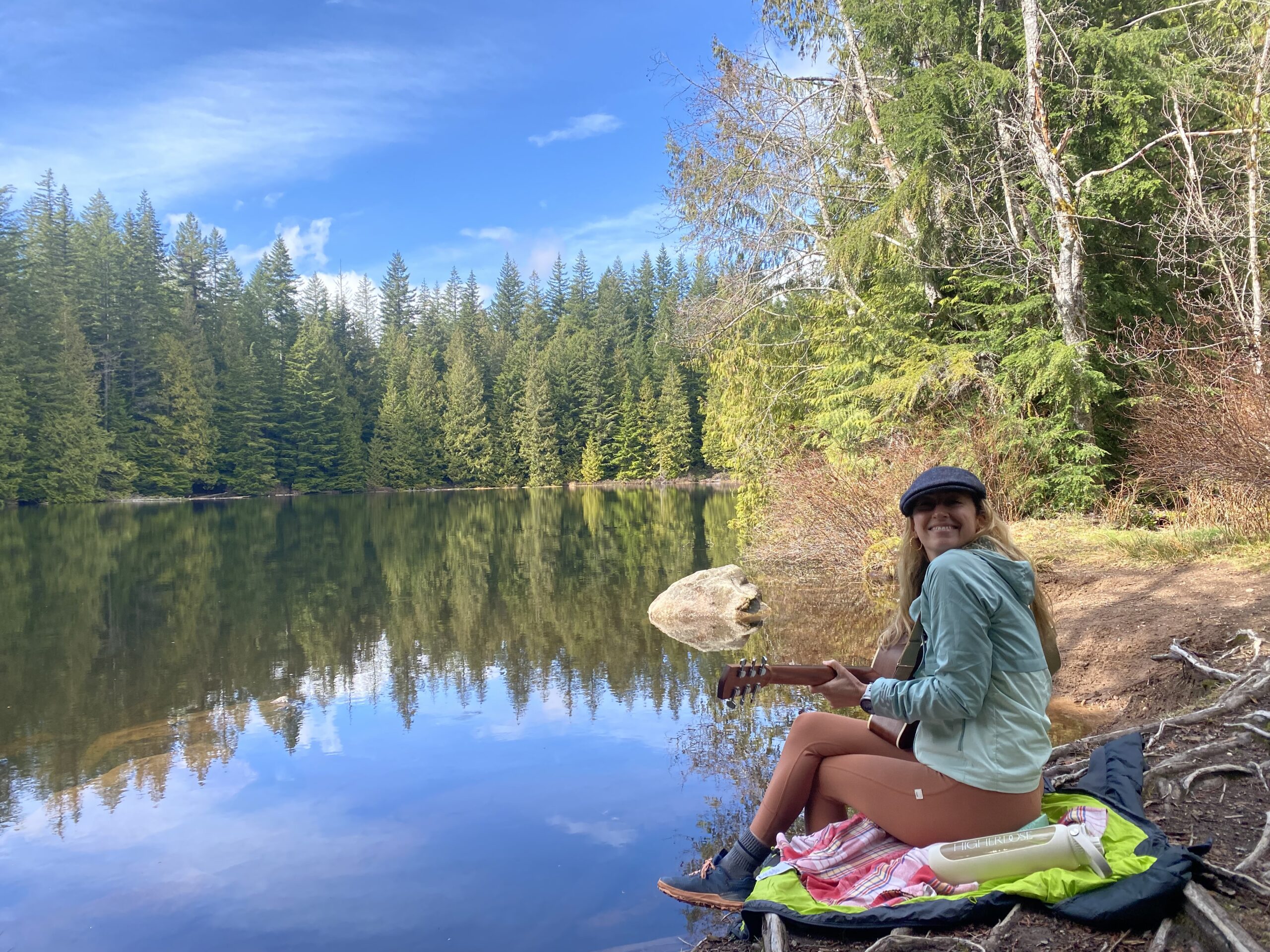 Woman sitting by a still alpine lake surrounded by pine trees, journaling on a sunny day in nature.