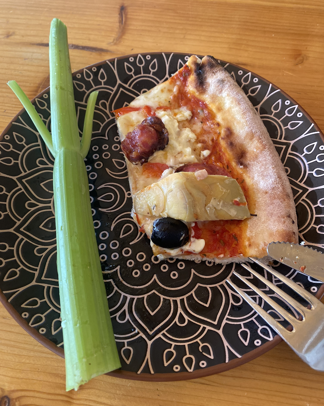 A slice of leftover pizza and a stick of celery on a patterned black plate, symbolizing the author’s reflection on food, culture, and adapting to life in the Swiss Alps.