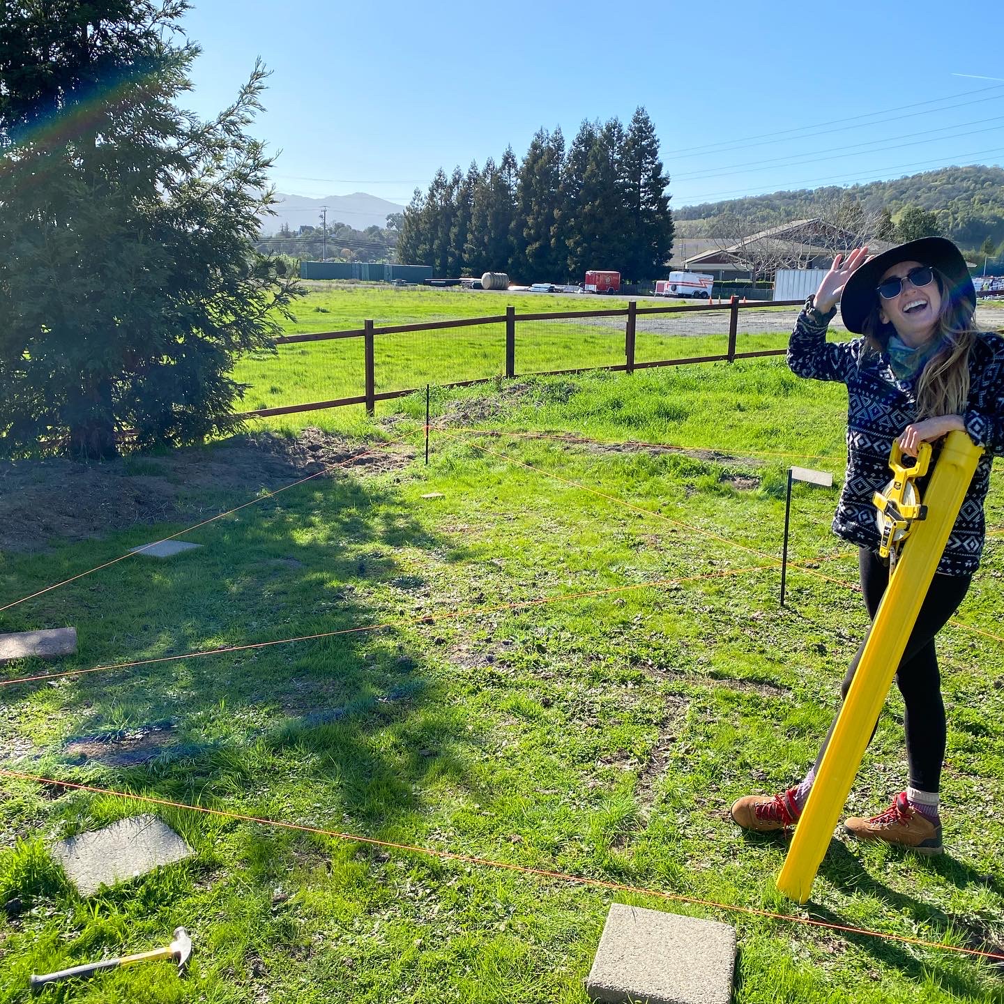 Woman standing on open grass holding a yellow measuring tool while string lines mark out the foundation of a future structure, representing the early stages of building something from the ground up.